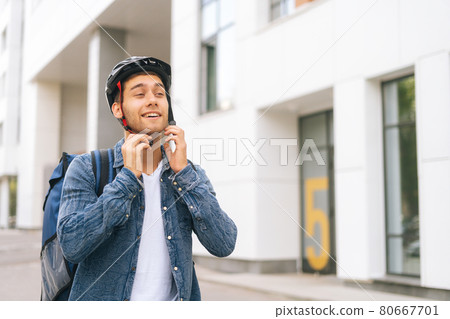 Front view of joyful young courier male with thermo backpack putting on helmet on head before to deliver an order to customer by bicycle. 80667701