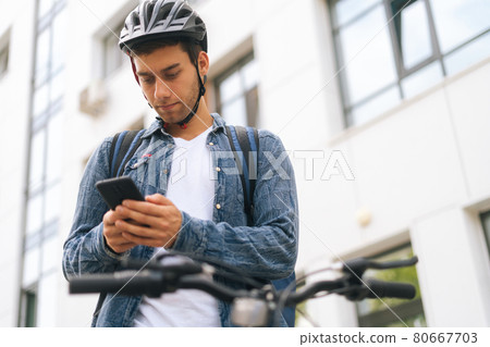 Low-angle view of handsome male courier with backpack standing with bicycle in city street and using navigation app on phone, close-up. Low-angle view of handsome male courier with backpack standing with bicycle in city street and using navigation app on phone, close-up. 80667703
