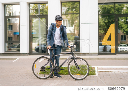Cheerful courier male getting out of client apartment, sitting on bicycle and riding off to next delivery. 80669979