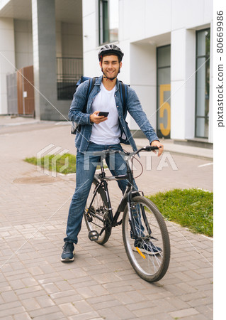 Full length vertical portrait of cheerful male courier with backpack standing with bicycle in city street and using navigation app on phone. 80669986