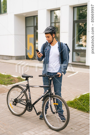 Full length portrait of handsome male courier with thermo backpack standing with bicycle in city street and using navigation app on phone. 80669997