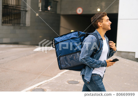 Side view of handsome male courier with thermo backpack walking in city street holding mobile phone is hand. Side view of handsome male courier with thermo backpack walking in city street holding mobile phone is hand. 80669999
