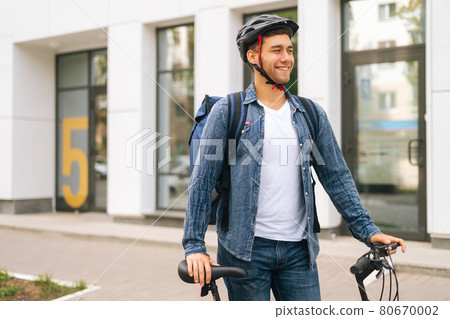Medium shot portrait of cheerful handsome young delivery man in protective helmet standing near bicycle in city street, looking away. 80670002