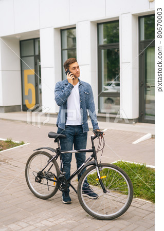 Vertical portrait of handsome young man in casual clothing standing with bicycle in city street and talking on mobile phone. Vertical portrait of handsome young man in casual clothing standing with bicycle in city street and talking on mobile phone. 80670003