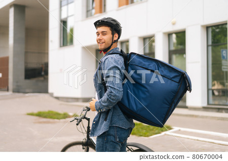Back view of smiling handsome young delivery man in protective helmet posing standing near bicycle in city street on background of office building Back view of smiling handsome young delivery man in protective helmet posing standing near bicycle in city street on background of office building 80670004