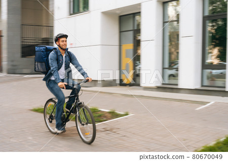 Cheerful handsome young delivery man with thermo backpack riding bicycle in city street on blurred background of office building. Cheerful handsome young delivery man with thermo backpack riding bicycle in city street on blurred background of office building. 80670049