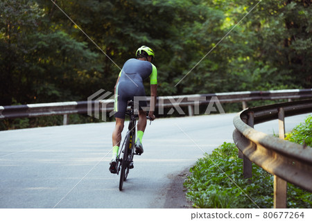 Young male triathlete riding a bicycle on the open road. Professional sportsman is engaged in triathlon on bright summer day Young male triathlete riding a bicycle on the open road. Professional sportsman is engaged in triathlon on bright summer day 80677264