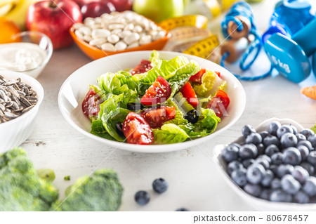 Healthy fresh salad with tomatoes olives and olive oil, surrounded by healthy food and exercise equipment Healthy fresh salad with tomatoes olives and olive oil, surrounded by healthy food and exercise equipment 80678457