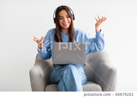 Happy young woman in headphones having online business meeting on laptop, sitting in comfy armchair on white background 80679281