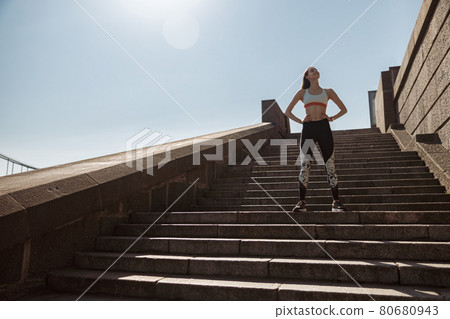 Woman in tracksuit with hands on waist stands on stone stairs on sunny summer day Woman in tracksuit with hands on waist stands on stone stairs on sunny summer day 80680943