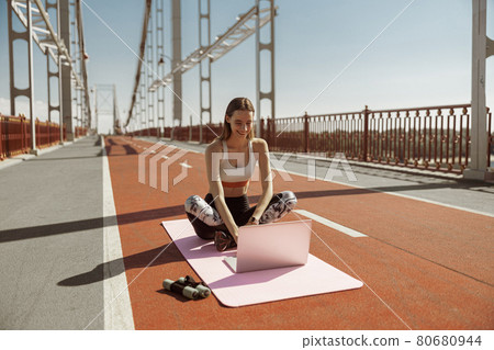 Cheerful woman watches video lesson via contemporary laptop sitting on footbridge Cheerful woman watches video lesson via contemporary laptop sitting on footbridge 80680944