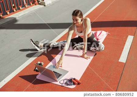 Positive lady with sports equipment chooses video tutorial for workout sitting on footbridge Positive lady with sports equipment chooses video tutorial for workout sitting on footbridge 80680952
