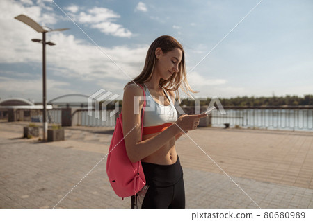 Positive woman in stylish tracksuit uses mobile phone standing on city waterfront Positive woman in stylish tracksuit uses mobile phone standing on city waterfront 80680989