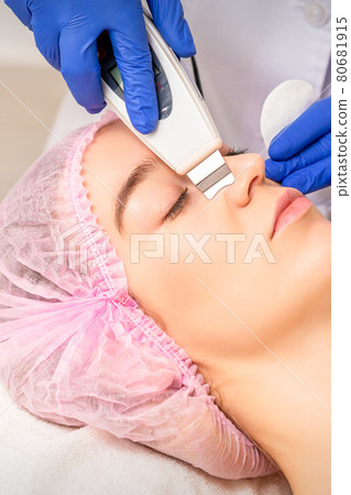 Closeup of beautiful young woman receiving ultrasound facial exfoliation and cavitation facial peeling with ultrasonic equipment in cosmetology office. Closeup of beautiful young woman receiving ultrasound facial exfoliation and cavitation facial peeling with ultrasonic equipment in cosmetology office. 80681915