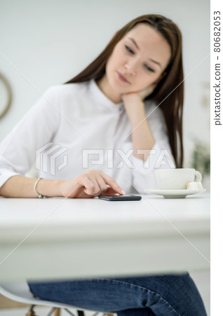 Young brunette in a white shirt is waiting for a business partner in a cafe. Woman at a business meeting. Girl drinks coffee during the break and uses a smartphone reads sms. Young brunette in a white shirt is waiting for a business partner in a cafe. Woman at a business meeting. Girl drinks coffee during the break and uses a smartphone reads sms. 80682053
