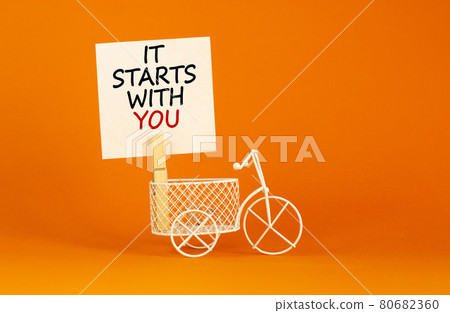 Wooden clothespin with white sheet of paper. Text 'it starts with you'. Miniature bicycle model. Beautiful orange background. Business concept, copy space. Wooden clothespin with white sheet of paper. Text 'it starts with you'. Miniature bicycle model. Beautiful orange background. Business concept, copy space. 80682360
