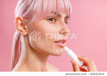 Close up portrait of beautiful young woman with pink hair looking aside, applying lip balm on her lips isolated over pink background 80682644