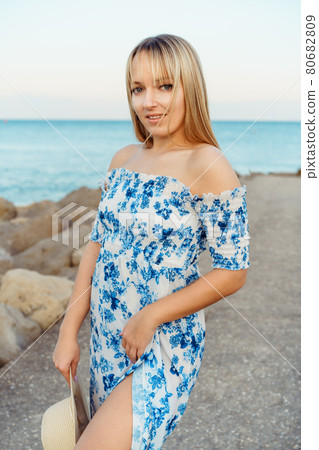 Portrait of a young blonde woman in summer dress with a straw hat on the seaside pier during a walk. Caucasian girl relaxing and enjoying peace on vacation. Vertical card, copy space. Portrait of a young blonde woman in summer dress with a straw hat on the seaside pier during a walk. Caucasian girl relaxing and enjoying peace on vacation. Vertical card, copy space. 80682809