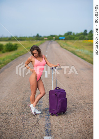 A beautiful brunette in a pink monokini is walking along a country road. Woman in a seductive swimsuit in the countryside with a luggage bag. 80683408