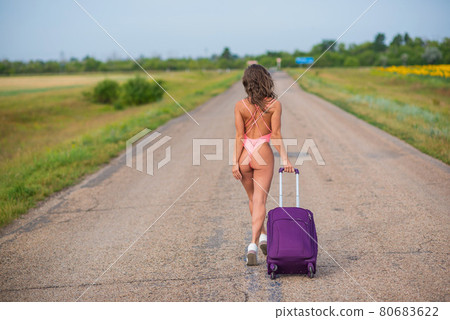 A beautiful brunette in a pink monokini is walking along a country road. Woman in a seductive swimsuit in the countryside with a luggage bag. 80683622