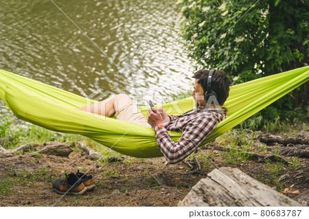 Outdoors and adventure concept. Bike trip to forest. Cyclist is resting in green hammock between trees in nature by lake while listening to music on headphones and surfing Internet on smartphone Outdoors and adventure concept. Bike trip to forest. Cyclist is resting in green hammock between trees in nature by lake while listening to music on headphones and surfing Internet on smartphone 80683787