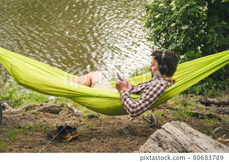 Young caucasian man resting in hammock, listening to music on headphones and using smartphone afterwards on bicycle in forest near the lake. Audio healing. Idyllic place. Travel, camping in nature Young caucasian man resting in hammock, listening to music on headphones and using smartphone afterwards on bicycle in forest near the lake. Audio healing. Idyllic place. Travel, camping in nature 80683789