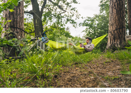 Man travels on bicycle, relaxing in green hammock, surfing Internet on smartphone, listening music on headphones in forest near lake. Cyclist in hammock at campsite by river. Male on bike in hammock Man travels on bicycle, relaxing in green hammock, surfing Internet on smartphone, listening music on headphones in forest near lake. Cyclist in hammock at campsite by river. Male on bike in hammock 80683790