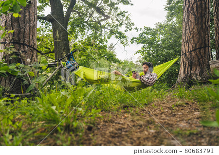 Man on bicycle trip at camping by lake is relaxing in green hammock while listening to music. Active recreation theme in nature. Hipster cyclist with headphones having fun in hammock by river Man on bicycle trip at camping by lake is relaxing in green hammock while listening to music. Active recreation theme in nature. Hipster cyclist with headphones having fun in hammock by river 80683791