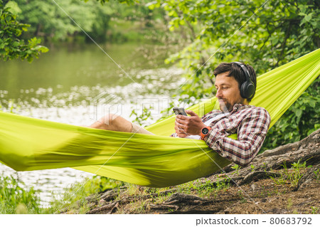Outdoors and adventure concept. Bike trip to forest. Cyclist is resting in green hammock between trees in nature by lake while listening to music on headphones and surfing Internet on smartphone Outdoors and adventure concept. Bike trip to forest. Cyclist is resting in green hammock between trees in nature by lake while listening to music on headphones and surfing Internet on smartphone 80683792