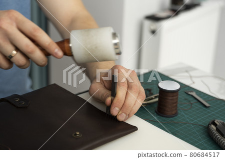 Mens hand holding a leather carving hammer and steel chisel hole punch and makes a leather wallet in his workshop. Working process with a brown natural leather. Craftsman holding a crafting tools. Mens hand holding a leather carving hammer and steel chisel hole punch and makes a leather wallet in his workshop. Working process with a brown natural leather. Craftsman holding a crafting tools. 80684517