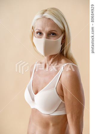 Portrait of beautiful caucasian mature blonde woman in underwear and protective face mask looking at camera while posing half naked against beige background Portrait of beautiful caucasian mature blonde woman in underwear and protective face mask looking at camera while posing half naked against beige background 80684929