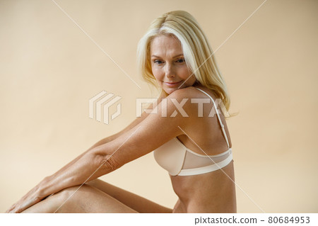Dreaming mature blonde woman in good shape posing in underwear, sitting on studio floor and looking away isolated over beige background Dreaming mature blonde woman in good shape posing in underwear, sitting on studio floor and looking away isolated over beige background 80684953
