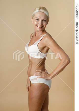 Pretty mature woman in underwear with fit body turning head to camera and smiling, holding hand on waist standing isolated over beige background Pretty mature woman in underwear with fit body turning head to camera and smiling, holding hand on waist standing isolated over beige background 80685449