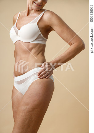 Cropped shot of confident mature woman in underwear with fit body smiling, holding hands on waist, posing, standing isolated over beige background Cropped shot of confident mature woman in underwear with fit body smiling, holding hands on waist, posing, standing isolated over beige background 80686062