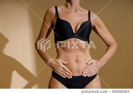 Cropped shot of perfect fit body of mature woman in black underwear holding hands on her stomach while posing half naked, standing isolated over beige background Cropped shot of perfect fit body of mature woman in black underwear holding hands on her stomach while posing half naked, standing isolated over beige background 80686065