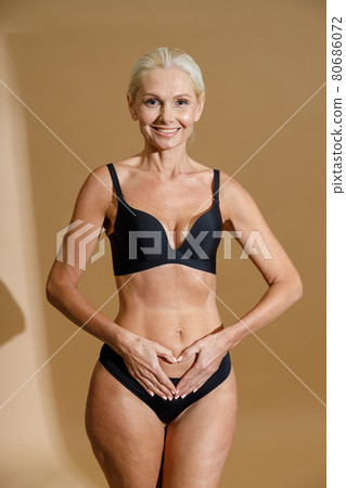 Studio shot of smiling mature blonde woman in black underwear holding hands on her belly, forming a heart symbol while standing isolated over beige background 80686072