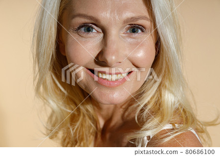Close up portrait of lovely caucasian mature blonde woman with natural makeup smiling at camera while posing isolated over beige background Close up portrait of lovely caucasian mature blonde woman with natural makeup smiling at camera while posing isolated over beige background 80686100