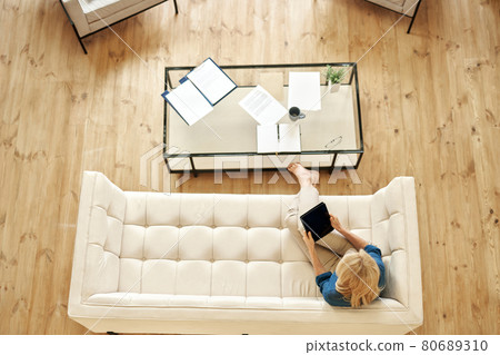 Top view of mature woman in casual wear using tablet pc while working from home, doing some paperwork, sitting on a couch in modern apartment 80689310