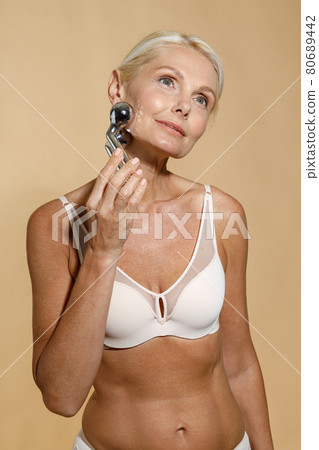 Beautiful mature blonde woman in underwear with glowing skin using metal face roller, looking away, posing isolated over beige background 80689442