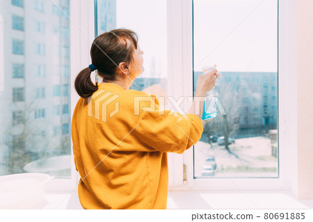 Woman Of Fifty In Yellow Sweater And Jeans Washes Dusty Window In Apartment. 50 Year Old Woman Cleans Windows From Stains Using Rag And Spray Cleaner. Caucasian Elderly Woman Is Cleaning House, Doing Woman Of Fifty In Yellow Sweater And Jeans Washes Dusty Window In Apartment. 50 Year Old Woman Cleans Windows From Stains Using Rag And Spray Cleaner. Caucasian Elderly Woman Is Cleaning House, Doing 80691885