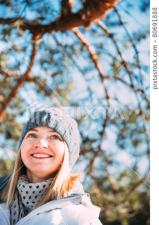 Young Caucasian Lady Woman Dressed In White Jacket And Grey-blue, Gray Hat Posing Near Pine Tree. Close Up Portrait. beautiful young woman smiling Young Caucasian Lady Woman Dressed In White Jacket And Grey-blue, Gray Hat Posing Near Pine Tree. Close Up Portrait. beautiful young woman smiling 80691888