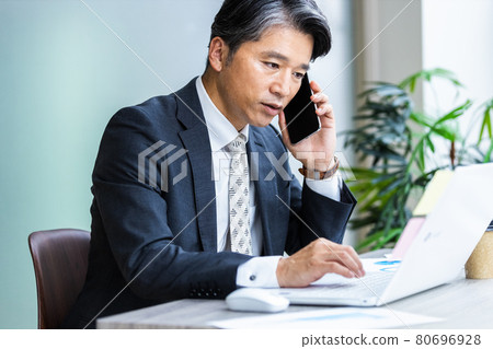 一個商人一邊看著電腦屏幕一邊用智能手機說話 80696928