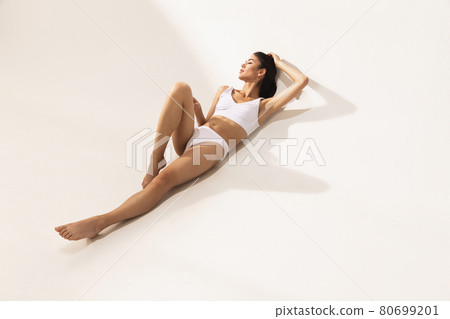 Portrait of young beautiful tanned woman in white underwear lying on floor over white background. Portrait of young beautiful tanned woman in white underwear lying on floor over white background. 80699201