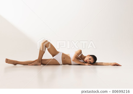 Portrait of young beautiful tanned woman in white underwear lying on floor over white background. Portrait of young beautiful tanned woman in white underwear lying on floor over white background. 80699216