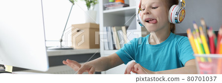 Little girl in headphones sitting at table and typing on computer keyboard Little girl in headphones sitting at table and typing on computer keyboard 80701037
