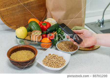 Whole-food, plant-based diet for boost health. Female hand cell phone with Plant-Based Menu recipes and healthy meal, vegetables, fruits, whole grains, healthy protein, oils. Vegan, vegetarian diet 80701200