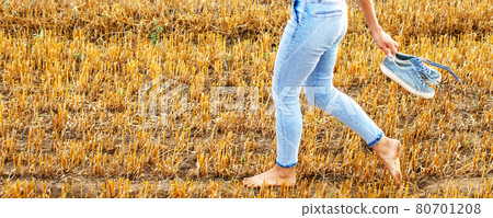 barefoot girl with sneakers in hand walking in the agricultural field with haystack and bales 80701208