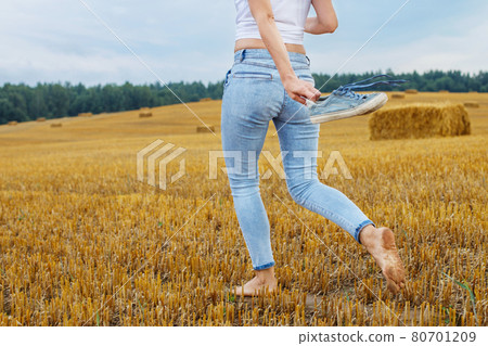 barefoot girl with sneakers in hand walking in the agricultural field with haystack and bales 80701209