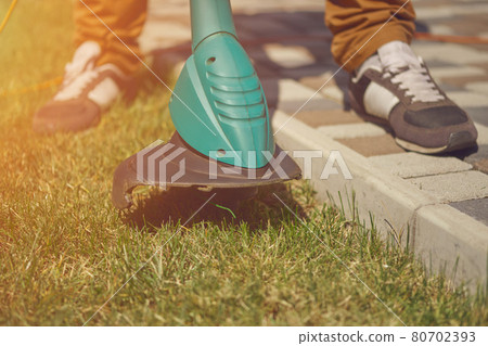 Male in sneakers and pants is cutting green grass with handheld modern lawn mower in his garden. Gardening care equipment and services. Close up Male in sneakers and pants is cutting green grass with handheld modern lawn mower in his garden. Gardening care equipment and services. Close up 80702393