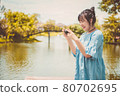 Asian woman in blue dress in public park carrying digital mirrorless camera and taking photo without facial mask in happy mood. People lifestyle and leisure concept. Outdoor travel and Nature theme. 80702695